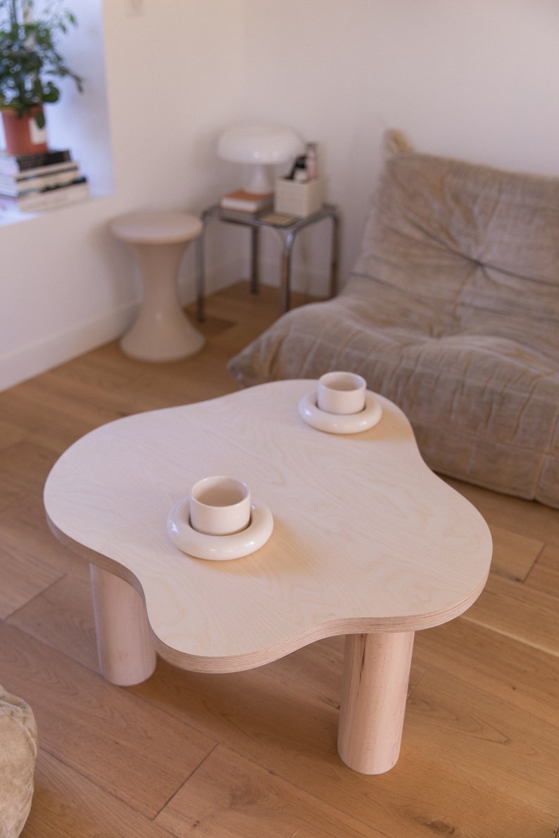 May include: A light wood coffee table with a unique cloud-shaped top and four rounded legs. Two white ceramic cups are placed on the table, each with a white saucer.