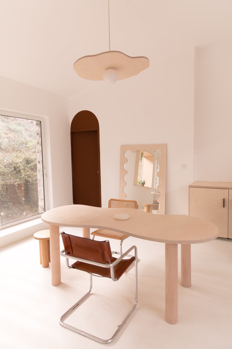 Puede incluir: Una mesa de madera clara con una forma org&aacute;nica &uacute;nica, con una silla de cuero marr&oacute;n y una l&aacute;mpara colgante blanca con una pantalla en forma de nube. La habitaci&oacute;n est&aacute; decorada en un estilo minimalista con paredes blancas y una gran ventana.