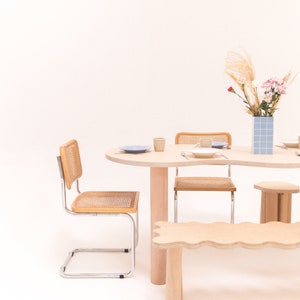 May include: A light wood dining table with a wavy edge and a light wood bench. Two chairs with woven cane seats and chrome frames are placed at the table. A vase with dried flowers and a blue and white tiled vase are on the table.