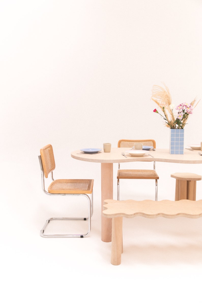 May include: A light wood dining table with a wavy edge and a light wood bench. Two chairs with woven cane seats and chrome frames are placed at the table. A vase with dried flowers and a blue and white tiled vase are on the table.