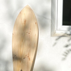 May include: A wooden surfboard with a natural finish. The surfboard is shaped like a fish and has a rounded nose.