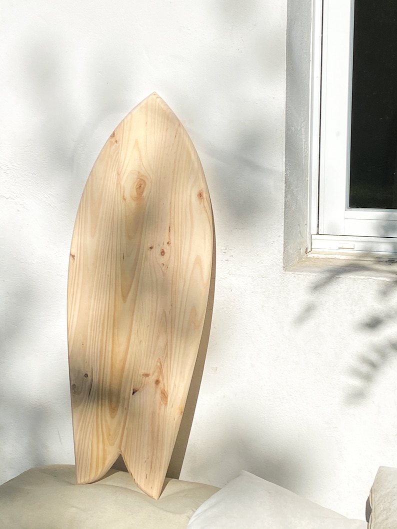 May include: A wooden surfboard with a natural finish. The surfboard is shaped like a fish and has a rounded nose.