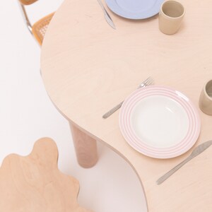 May include: A light wood table with a wavy edge and two chairs. The table is set with two place settings, each with a plate, a fork, a knife, and a mug.