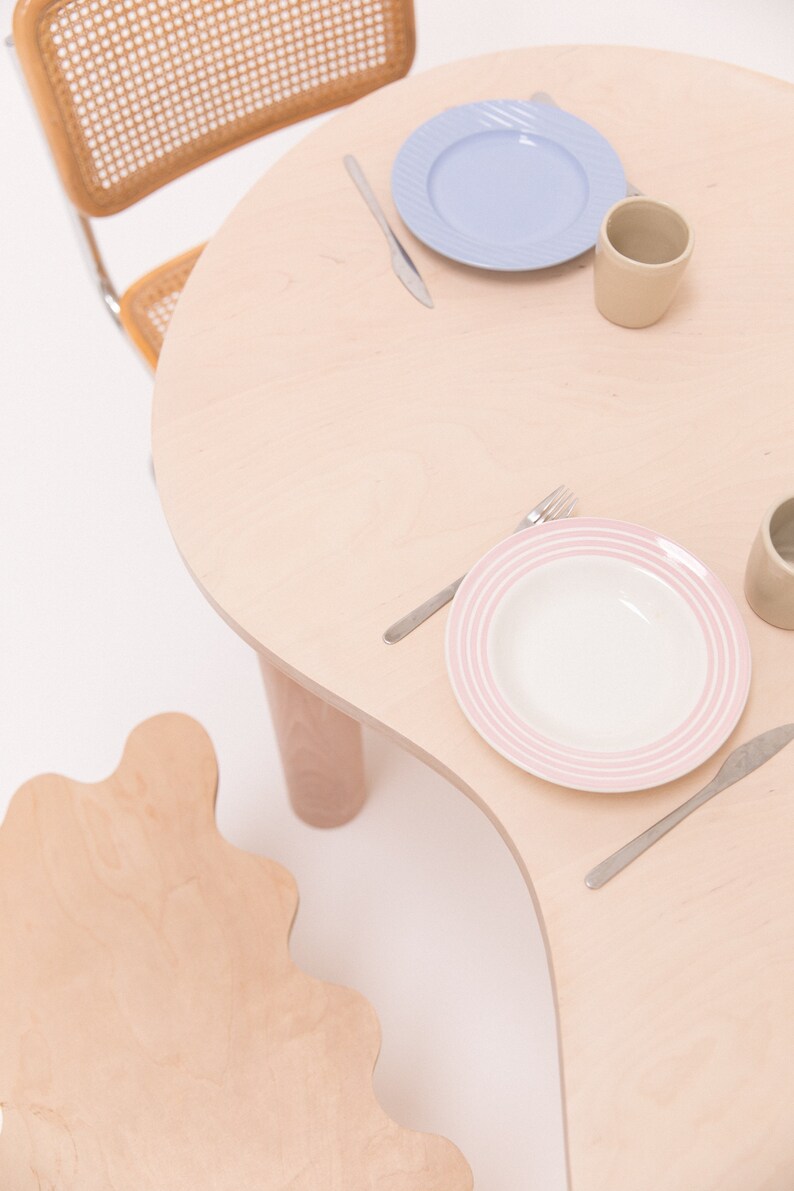 May include: A light wood table with a wavy edge and two chairs. The table is set with two place settings, each with a plate, a fork, a knife, and a mug.