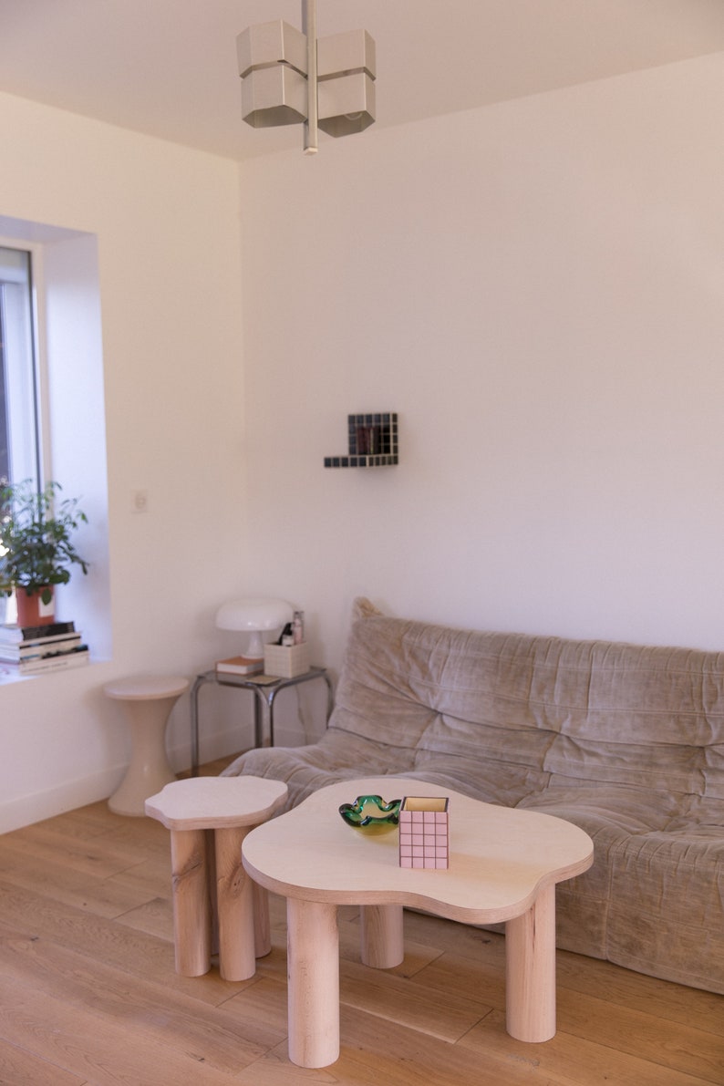 May include: Two wooden coffee tables with irregular cloud-like tops. The larger table has a pink and yellow patterned box and a green glass bowl on top. The smaller table is next to the larger table. Both tables have thick wooden legs.
