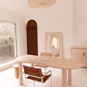 May include: A light wood table with a unique, organic shape, featuring a brown leather chair and a woven wicker chair. The table has four light wood legs and is set against a white wall with a large, organic-shaped mirror and a light wood cabinet.