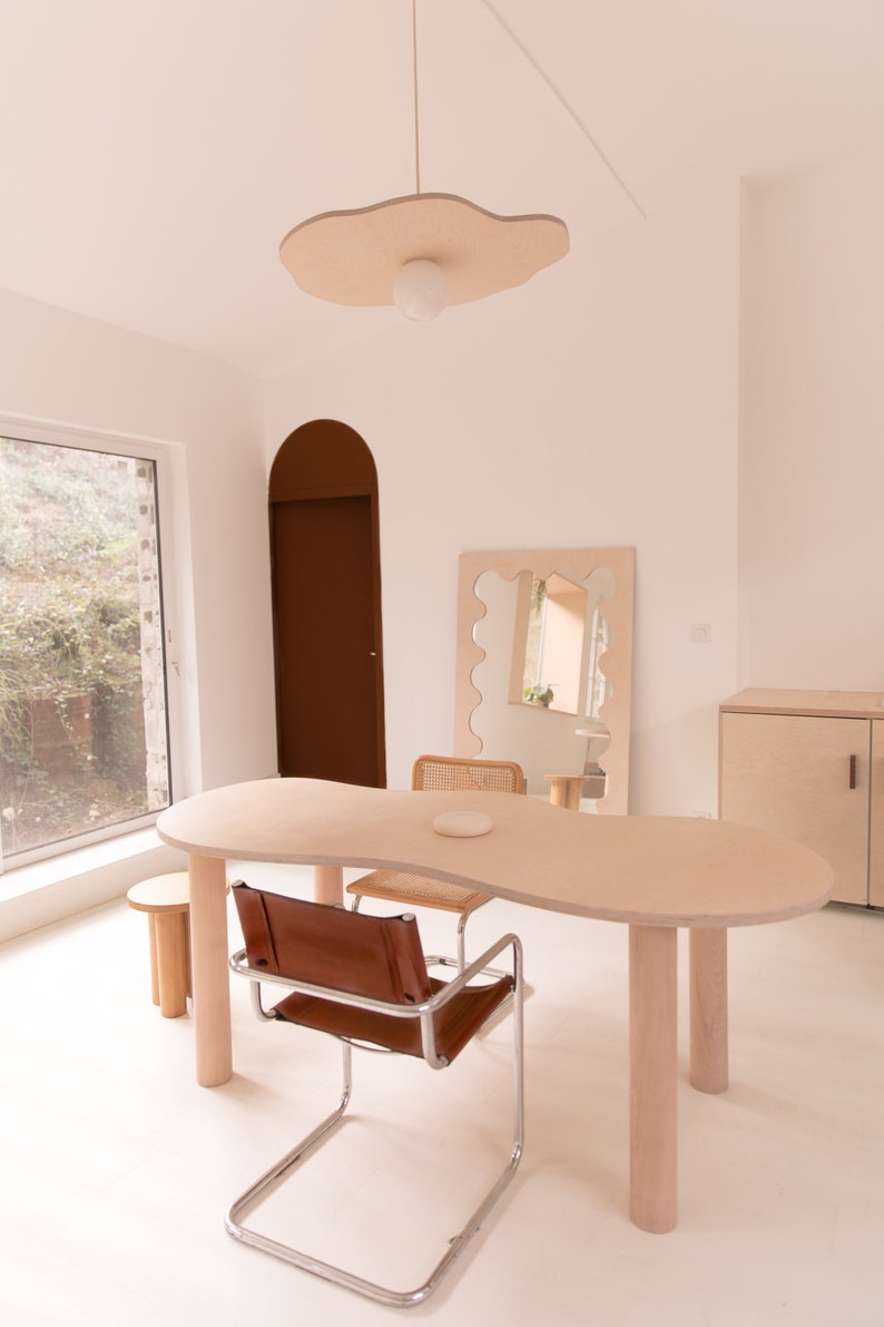 May include: A light wood table with a unique, organic shape, featuring a brown leather chair and a woven wicker chair. The table has four light wood legs and is set against a white wall with a large, organic-shaped mirror and a light wood cabinet.