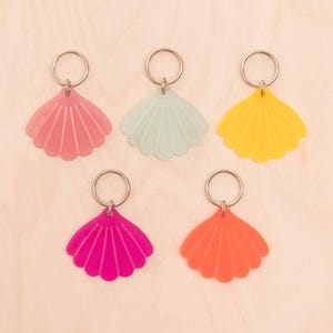 May include: Five seashell-shaped keyrings in pink, mint green, yellow, hot pink, and orange. Each keyring has a silver-coloured ring attached at the top. The keyrings are arranged on a light wood grain surface.