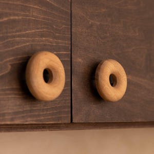 Peut inclure: Gros plan sur deux boutons de placard en bois en forme de donut. Les boutons sont brun clair et montés sur une porte d'armoire brun foncé. Le grain du bois est visible sur la porte et les boutons.