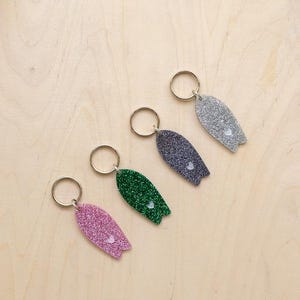 Op de afbeelding: Vier glitterende surfplank sleutelhangers in roze, groen, grijs en zilver, elk met een klein hartje. De sleutelhangers zijn gerangschikt op een licht houten oppervlak, en bieden een speels en kleurrijk accessoire.