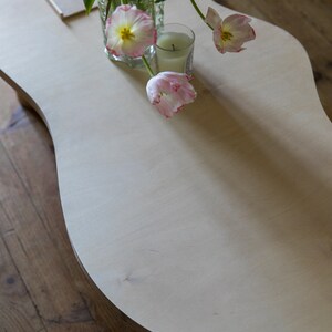 May include: A white, organic-shaped coffee table with a light wood finish. A bouquet of pink tulips in a clear glass vase sits on the table, along with a white candle.