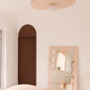 Puede incluir: Una mesa de madera clara con un borde ondulado y un objeto redondo blanco en la parte superior. Un espejo con un borde ondulado est&aacute; en el fondo, reflejando una habitaci&oacute;n con una puerta marr&oacute;n y una ventana. Una l&aacute;mpara con un globo blanco cuelga del techo.