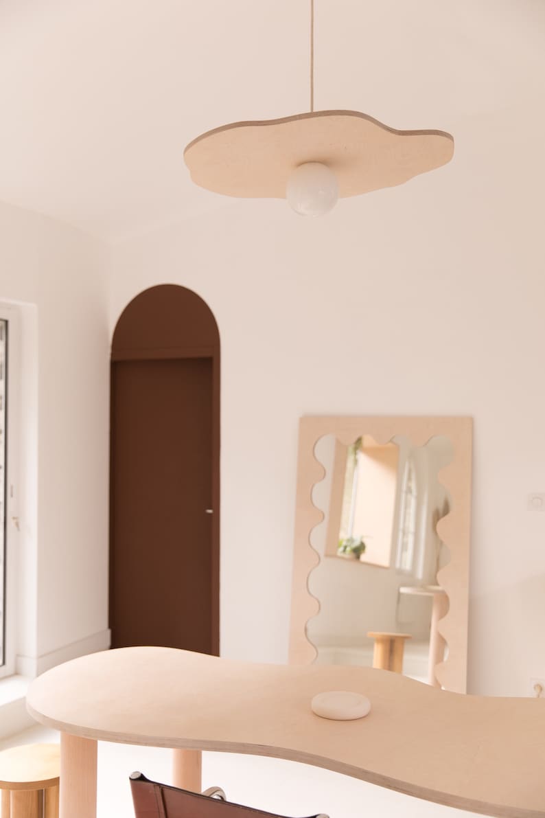 Puede incluir: Una mesa de madera clara con un borde ondulado y un objeto redondo blanco en la parte superior. Un espejo con un borde ondulado est&aacute; en el fondo, reflejando una habitaci&oacute;n con una puerta marr&oacute;n y una ventana. Una l&aacute;mpara con un globo blanco cuelga del techo.