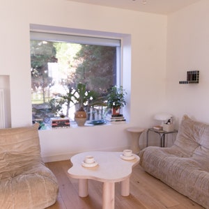 May include: A light wood coffee table with a unique cloud-shaped top and four cylindrical legs. Two white ceramic mugs are on the table, with a window and a couch in the background.