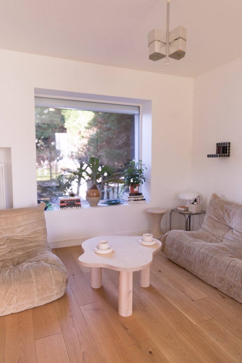 May include: A light wood coffee table with a unique cloud-shaped top and four cylindrical legs. Two white ceramic mugs are on the table, with a window and a couch in the background.