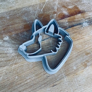 Puede incluir: Un cortador de galletas de plástico gris con forma de cabeza de perro, con orejas, hocico y pelaje detallados. El cortador está diseñado para crear galletas o golosinas con forma de perro. El cortador está sobre una superficie de madera.