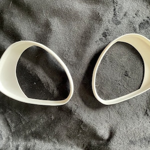 Peut inclure: Deux caches de phares de voiture en plastique blanc, conçus pour protéger les phares des dommages. Les caches sont en forme de croissant de lune et sont posés sur une surface en tissu noir.