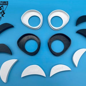 Könnte beinhalten: Ein Satz schwarzer und weißer Plastikteile zum Basteln. Die Teile sind in Form von Augen, Augenbrauen und Nasen. Die Teile sind auf einem blauen Hintergrund angeordnet.