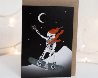 May include: Greeting card featuring a skeleton snowboarding under a crescent moon and stars. The skeleton wears a red Santa hat and scarf. The background is black with white mountains. The card is inside a brown envelope.
