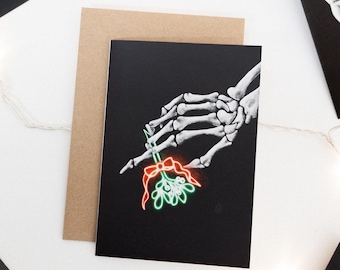 May include: A black card with a white skeleton hand holding a sprig of mistletoe with a red bow. The mistletoe is glowing green and the bow is glowing red.