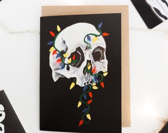 May include: A black card with a white skull with colorful Christmas lights wrapped around it. The skull is painted in a realistic style.