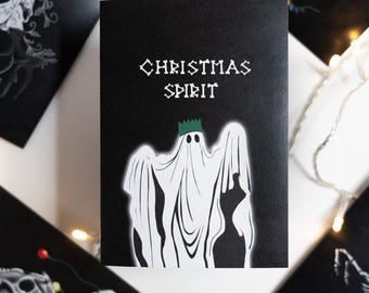 May include: Black greeting card with the words "CHRISTMAS SPIRIT" and a ghost wearing a crown. Other cards with skull and antler designs are scattered around the main card. Fairy lights are in the background.
