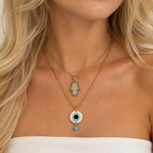 Puede incluir: Un collar de capas en tono dorado con dos colgantes distintos. El colgante superior es un dije de mano de Hamsa con detalles en azul y blanco. El colgante inferior es un dije circular de ojo turco con base blanca, centro negro y una peque&ntilde;a cuenta azul.