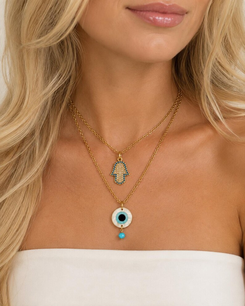 Puede incluir: Un collar de capas en tono dorado con dos colgantes distintos. El colgante superior es un dije de mano de Hamsa con detalles en azul y blanco. El colgante inferior es un dije circular de ojo turco con base blanca, centro negro y una peque&ntilde;a cuenta azul.