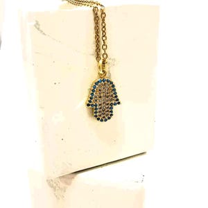 Puede incluir: Un collar de tono dorado con un colgante de mano de Hamsa. El colgante est&aacute; adornado con un grupo central de piedras claras y un borde de peque&ntilde;as piedras azules. La cadena es un modelo de eslabones finos en tono dorado, de aspecto cl&aacute;sico.
