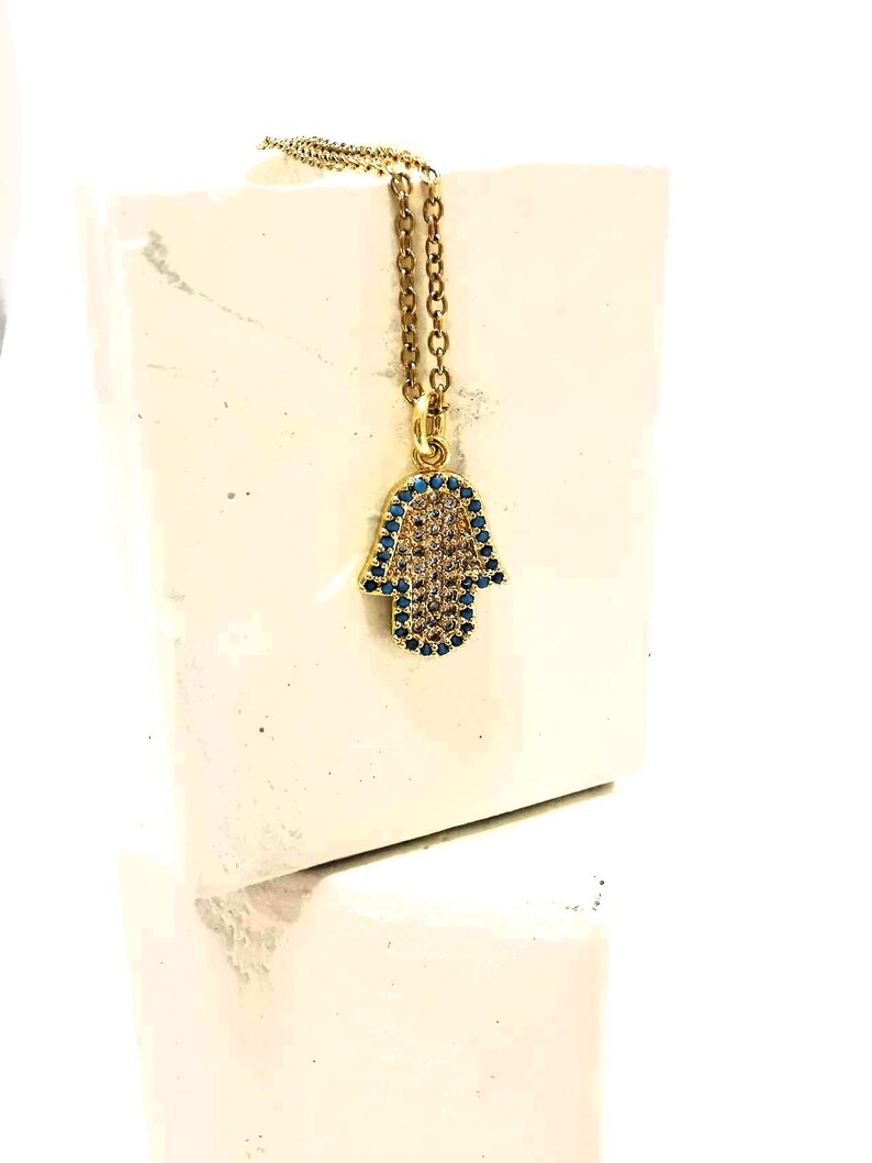 Puede incluir: Un collar de tono dorado con un colgante de mano de Hamsa. El colgante est&aacute; adornado con un grupo central de piedras claras y un borde de peque&ntilde;as piedras azules. La cadena es un modelo de eslabones finos en tono dorado, de aspecto cl&aacute;sico.