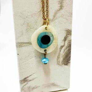 Puede incluir: Un collar de cadena en tono dorado con un colgante circular que presenta una base de concha blanca, una incrustaci&oacute;n en forma de ojo azul y negro, y una peque&ntilde;a cuenta facetada de color azul claro que cuelga de la parte inferior del colgante.