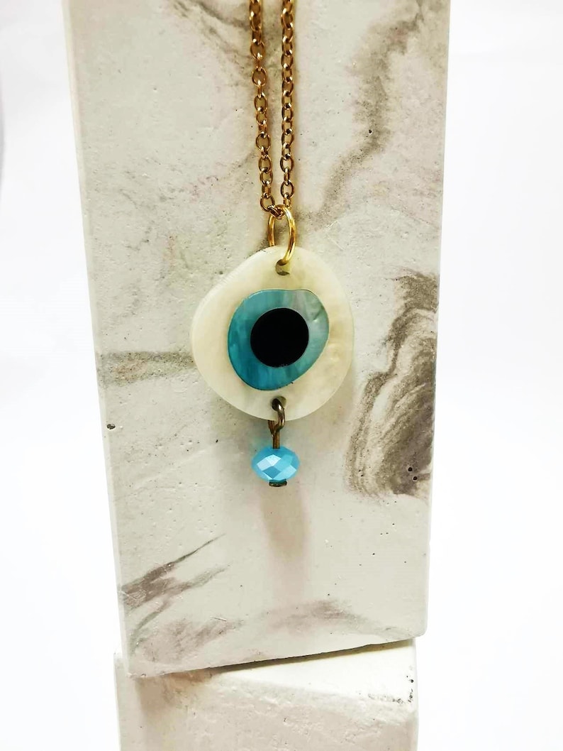 Puede incluir: Un collar de cadena en tono dorado con un colgante circular que presenta una base de concha blanca, una incrustaci&oacute;n en forma de ojo azul y negro, y una peque&ntilde;a cuenta facetada de color azul claro que cuelga de la parte inferior del colgante.