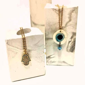 Puede incluir: Dos collares de cadena dorada. Uno presenta un colgante de mano de Hamsa dorado incrustado con peque&ntilde;as piedras azules. El otro collar tiene un colgante blanco circular con un dise&ntilde;o de ojo azul central y una peque&ntilde;a cuenta azul claro colgante.