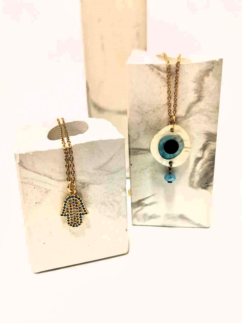 Puede incluir: Dos collares de cadena dorada. Uno presenta un colgante de mano de Hamsa dorado incrustado con peque&ntilde;as piedras azules. El otro collar tiene un colgante blanco circular con un dise&ntilde;o de ojo azul central y una peque&ntilde;a cuenta azul claro colgante.