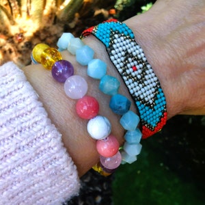 Può includere: Un set di braccialetti sul polso di una persona. Il braccialetto inferiore è fatto di perline di pietre preziose colorate. Il braccialetto superiore è un design intrecciato con perline turchesi, bianche, nere e rosse.