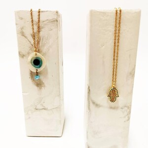 Puede incluir: Dos collares de cadena dorada expuestos sobre soportes rectangulares con patr&oacute;n de m&aacute;rmol blanco. Un collar presenta un colgante circular de ojo turco con centro azul y una peque&ntilde;a cuenta colgante. El otro collar tiene un colgante de mano de F&aacute;tima.