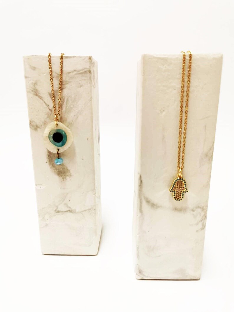 Puede incluir: Dos collares de cadena dorada expuestos sobre soportes rectangulares con patr&oacute;n de m&aacute;rmol blanco. Un collar presenta un colgante circular de ojo turco con centro azul y una peque&ntilde;a cuenta colgante. El otro collar tiene un colgante de mano de F&aacute;tima.