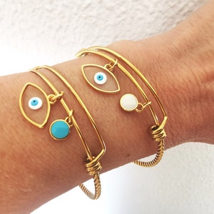 May include: Two gold bangle bracelets with evil eye charms. One charm has a white eye with a blue pupil and a turquoise stone. The other charm has a white eye with a blue pupil and a white stone.
