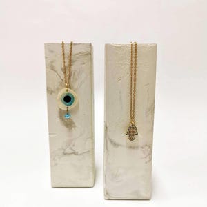 Puede incluir: Dos collares dorados exhibidos en soportes rectangulares con patr&oacute;n de m&aacute;rmol. Un collar presenta un colgante circular de ojo turco con un centro azul y una peque&ntilde;a cuenta colgante. El otro collar tiene un colgante de mano de Hamsa con detalles azules.