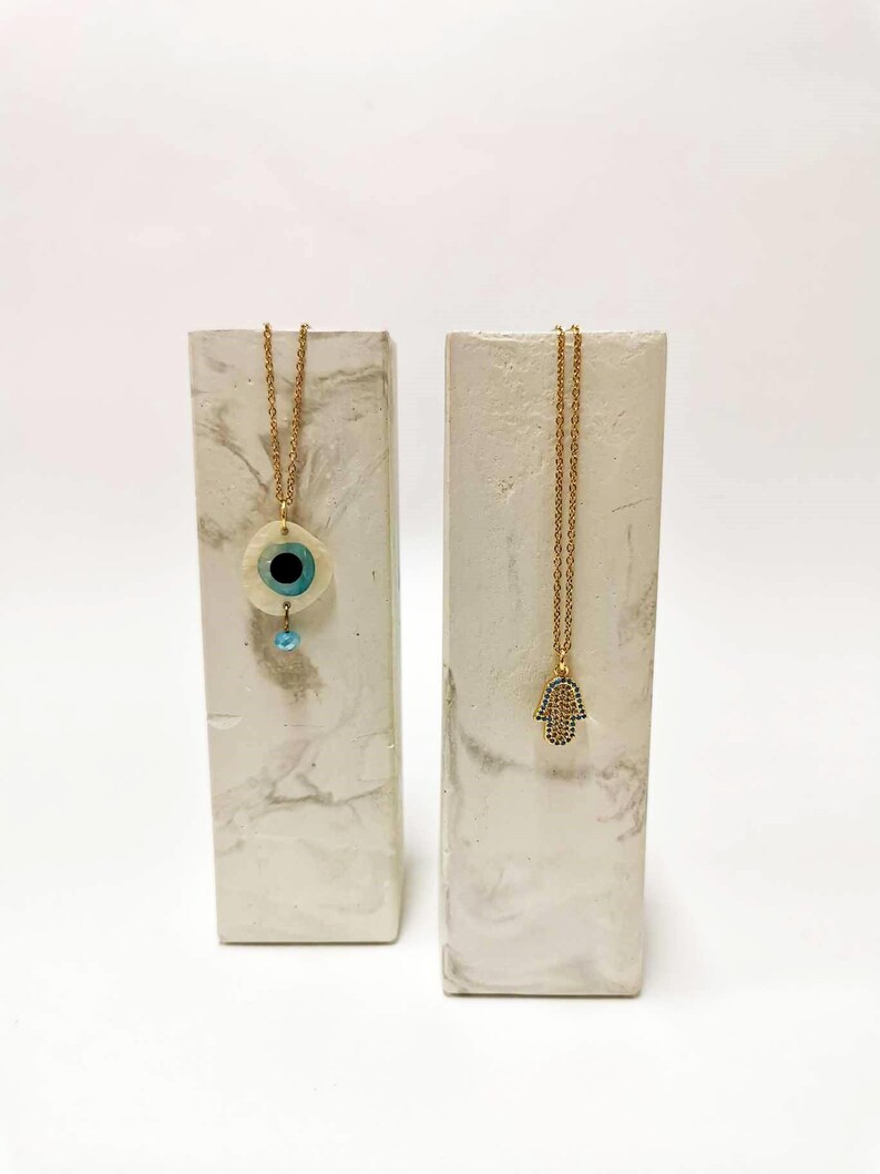 Puede incluir: Dos collares dorados exhibidos en soportes rectangulares con patr&oacute;n de m&aacute;rmol. Un collar presenta un colgante circular de ojo turco con un centro azul y una peque&ntilde;a cuenta colgante. El otro collar tiene un colgante de mano de Hamsa con detalles azules.