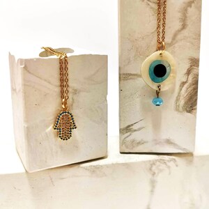 Puede incluir: Dos collares dorados. Uno presenta un colgante de mano de Hamsa incrustado con peque&ntilde;as piedras azules y transparentes. El otro collar tiene un colgante circular con una base blanca nacarada, un dise&ntilde;o de ojo azul y negro y una cuenta azul.