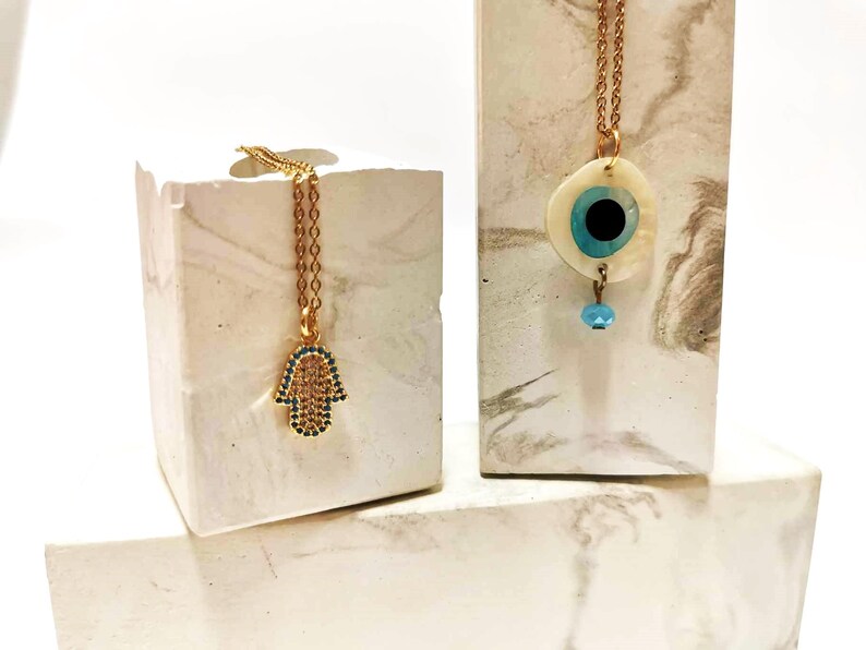 Puede incluir: Dos collares dorados. Uno presenta un colgante de mano de Hamsa incrustado con peque&ntilde;as piedras azules y transparentes. El otro collar tiene un colgante circular con una base blanca nacarada, un dise&ntilde;o de ojo azul y negro y una cuenta azul.