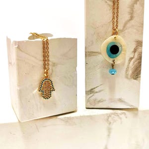 Puede incluir: Dos collares dorados. Uno presenta un colgante de mano de Hamsa incrustado con peque&ntilde;as piedras azules y transparentes. El otro collar tiene un colgante circular de color hueso con un dise&ntilde;o de ojo azul y negro y una peque&ntilde;a cuenta azul.