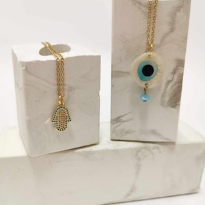 Puede incluir: Dos collares de cadena dorada. Uno presenta un colgante de mano de Hamsa incrustado con peque&ntilde;as piedras claras y azules. El otro presenta un colgante de concha blanca circular con un dise&ntilde;o de ojo azul y negro y una peque&ntilde;a cuenta azul.