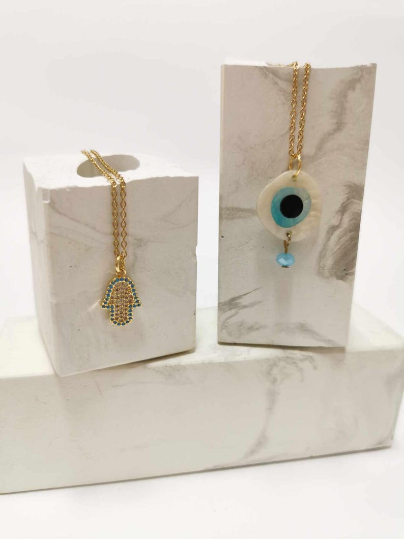 Puede incluir: Dos collares de cadena dorada. Uno presenta un colgante de mano de Hamsa incrustado con peque&ntilde;as piedras claras y azules. El otro presenta un colgante de concha blanca circular con un dise&ntilde;o de ojo azul y negro y una peque&ntilde;a cuenta azul.