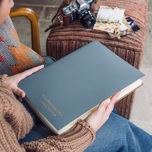 a woman is holding an A4 sized grey leather album which has been personalised with names on thr front. There is a camera on the side and some wedding photos. She is wearing jeans and a jumper