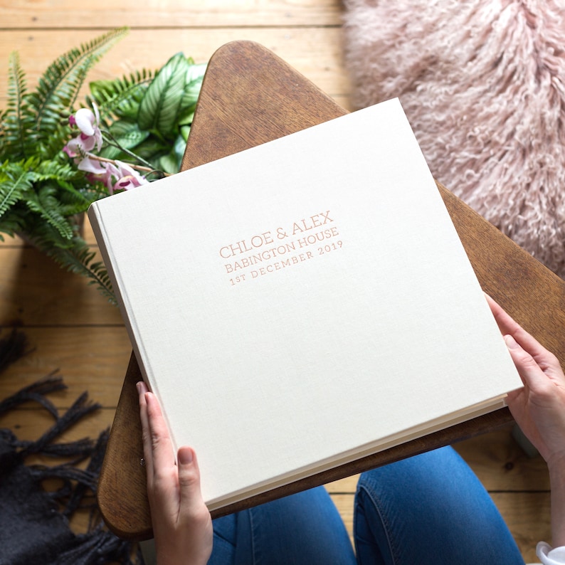 a large wedding photo album made from linen bookcloth in someones hands as they are about to look through it. Placed on a wooden table on top of a pink rug