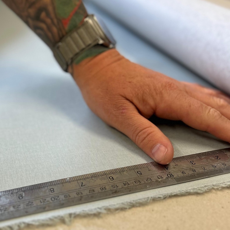A man is holding a metal ruler against a piece of blue linen cloth. He is measuring it. His hand is covered in tattoos.