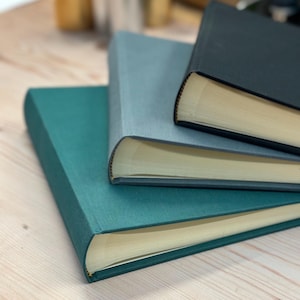Three linen photo albums are piled up on top of each other - a dark green one on the bottom, a mid grey one in the middle and then a black one on the top. You can see the spines and the pages inside.