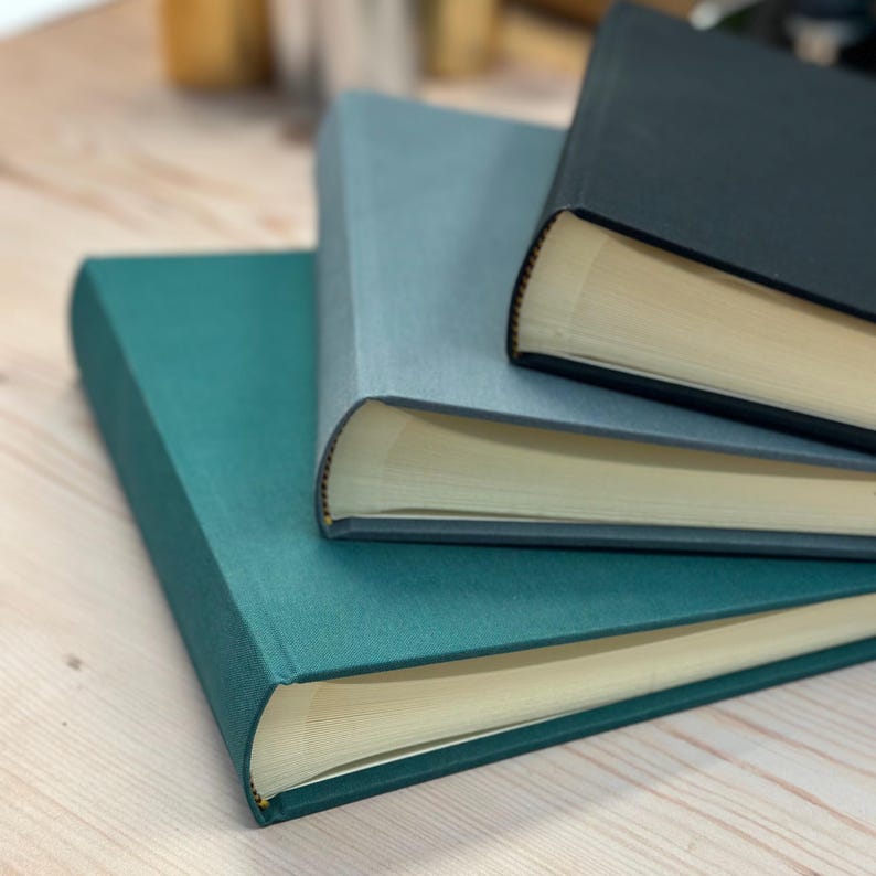 Three linen photo albums are piled up on top of each other - a dark green one on the bottom, a mid grey one in the middle and then a black one on the top. You can see the spines and the pages inside.